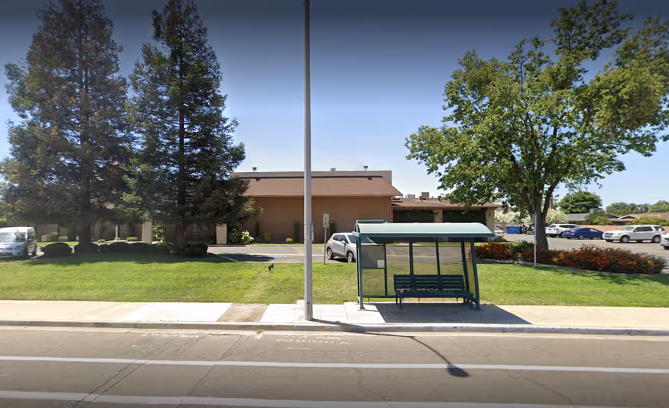 Street-facing view of an assisted living building with a green bus shelter in front, a lawn, trees, and parked cars.