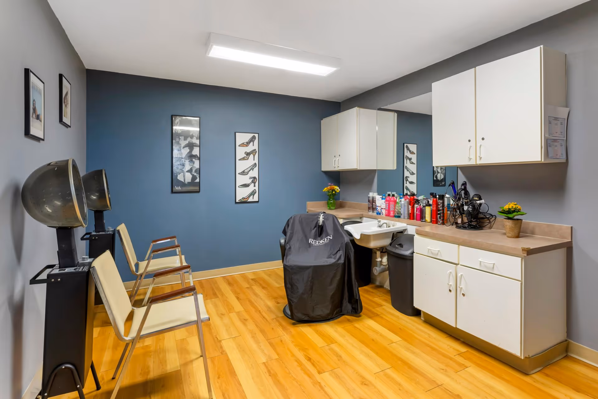 A small interior salon room with hooded hair dryers, styling chairs, a covered salon chair by a sink, and cabinets.