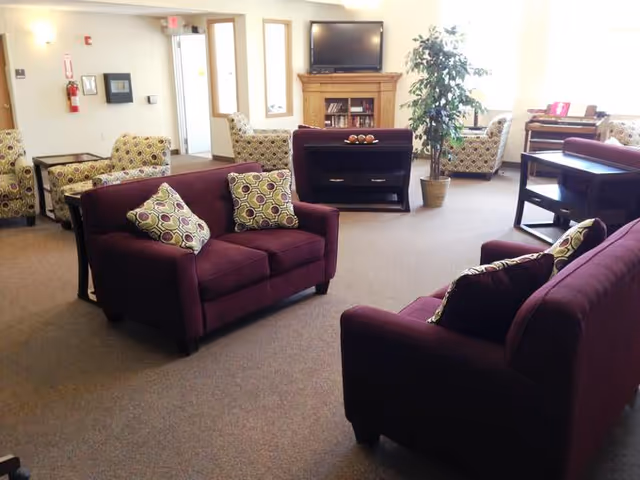 A well-lit living room area with two maroon sofas adorned with patterned cushions, several armchairs with a circular pattern, a wooden TV stand with a flat-screen television, a fireplace, a potted plant, and a piano in the background near a window.