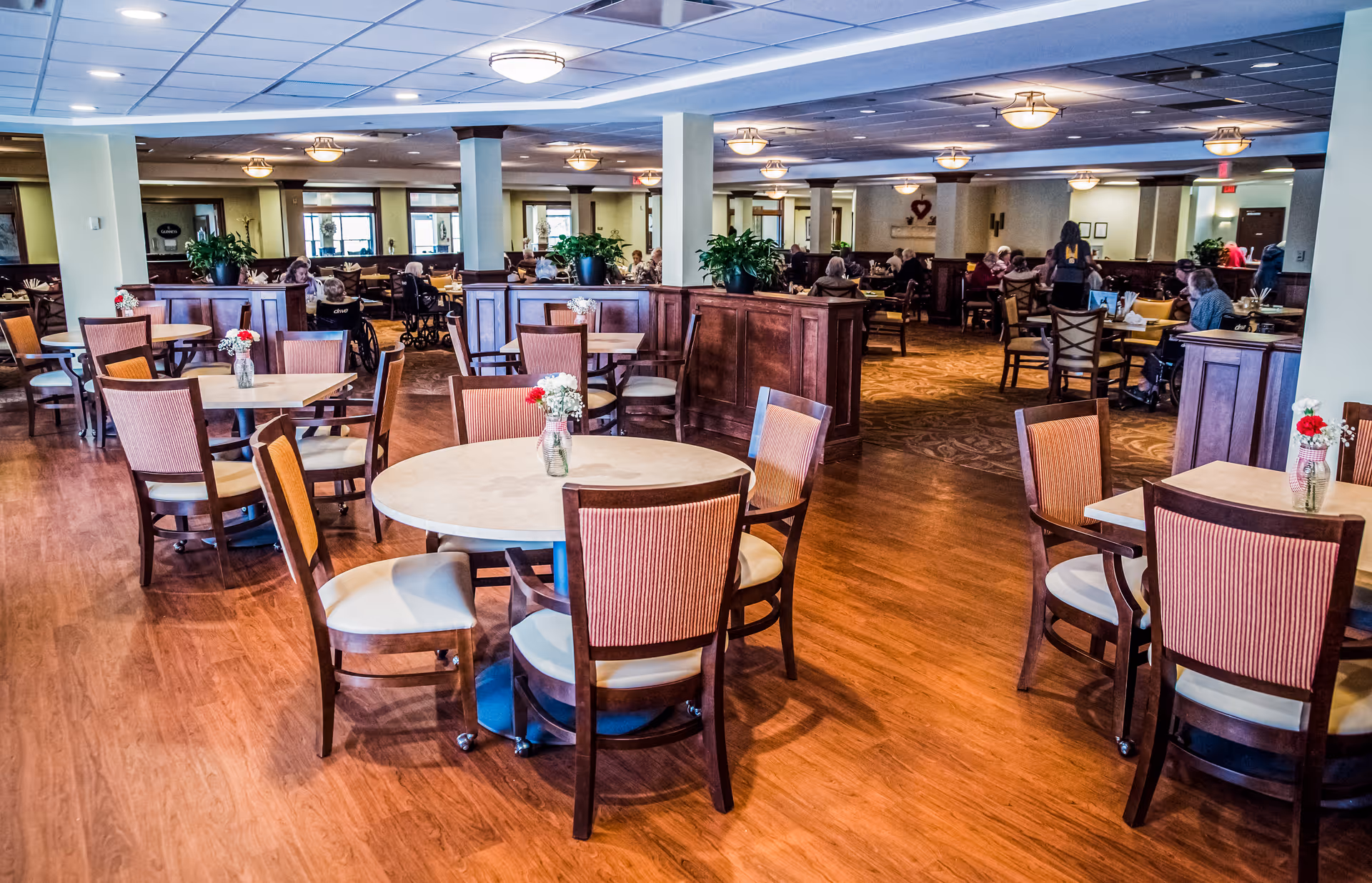 Spacious communal dining room with round and square tables, wooden chairs, floral centerpieces, and residents seated in the background.