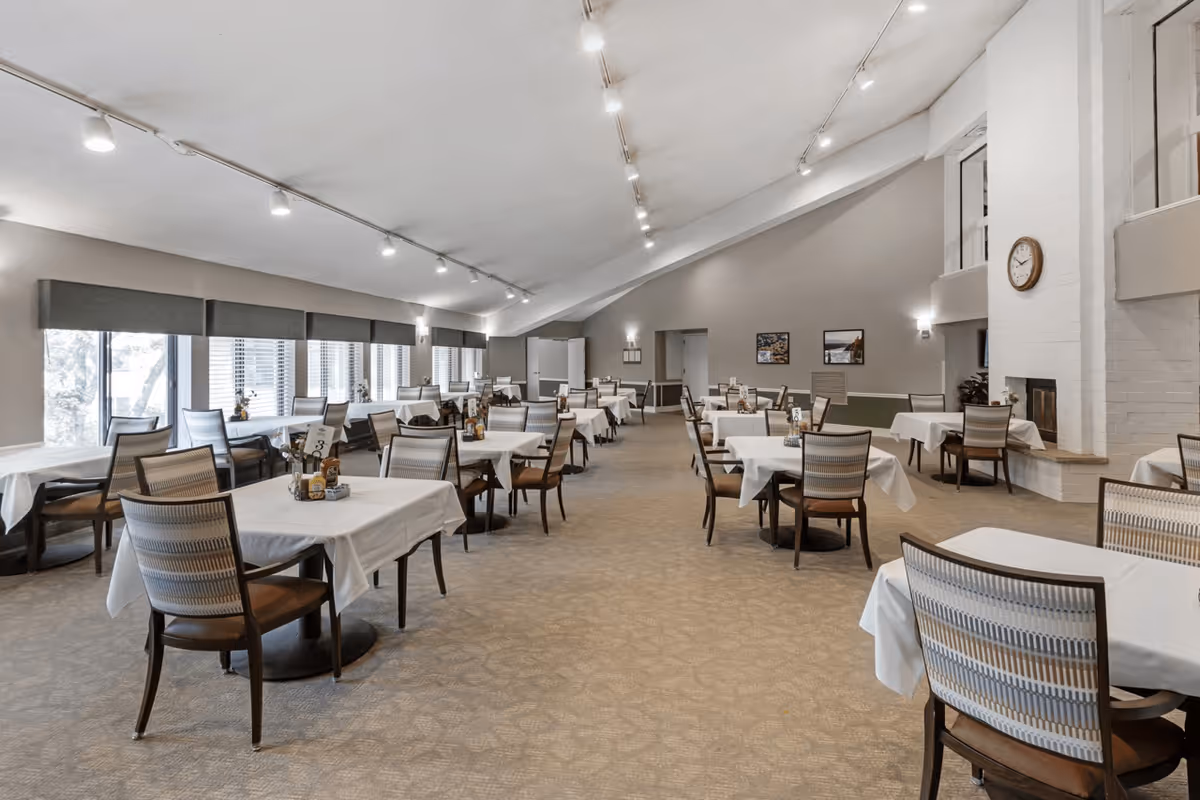 A spacious dining room with multiple tables covered in white tablecloths, each surrounded by wooden chairs with patterned cushions. Large windows on the left side allow natural light to fill the room. The walls are painted gray, and there is a white brick fireplace with a clock above it on the right side. The ceiling has track lighting, and the room has a calm, inviting atmosphere.