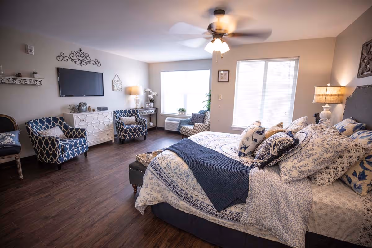 A spacious bedroom with a large bed covered in multiple patterned pillows and a blue throw blanket. The room has wooden flooring, two large windows with blinds, a ceiling fan with lights, two patterned armchairs, a white dresser with decorative items, a wall-mounted TV, and a side table with a lamp and flowers.