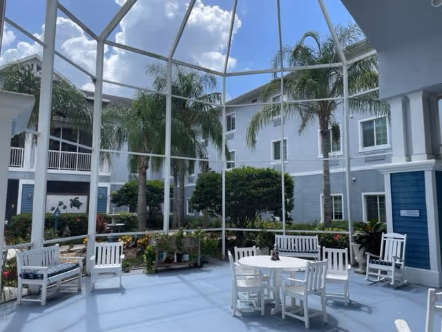 A bright, enclosed outdoor patio area with white wooden chairs and a round table. The space is surrounded by a white metal frame with a clear roof, allowing a view of blue sky and clouds. Palm trees and green shrubs are visible inside the enclosure, with a multi-story building in the background.