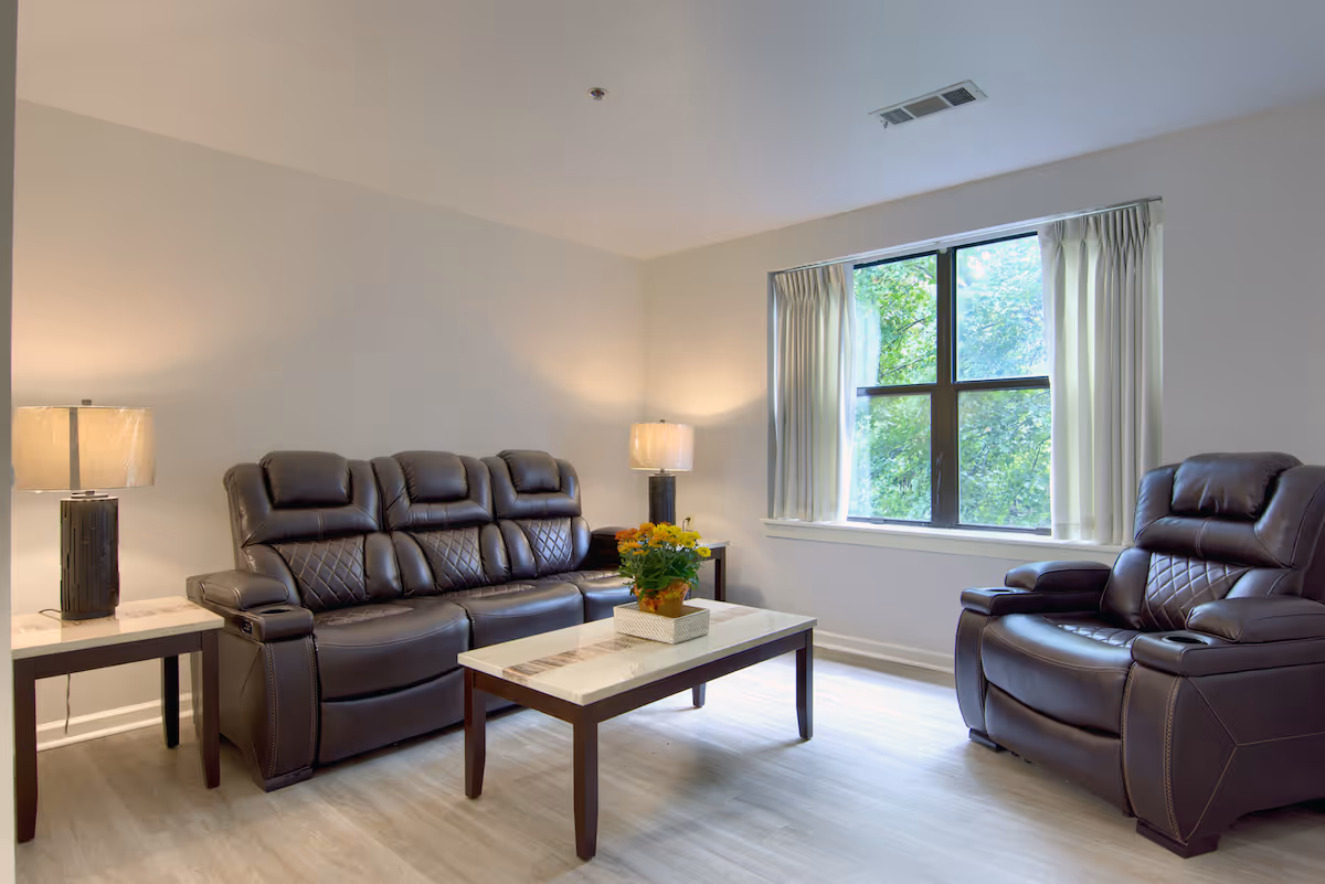 A bright seating area with a dark leather sofa and matching recliner around a central coffee table beneath a window.