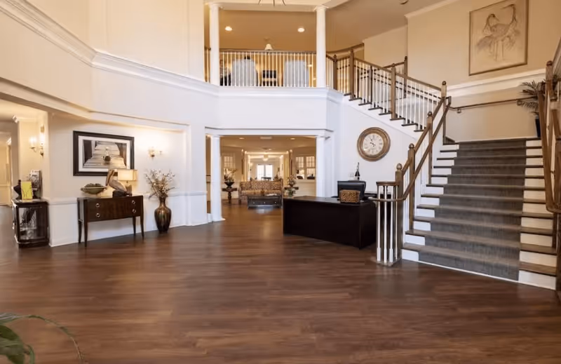 Spacious and elegant interior lobby area with wooden flooring, a staircase with carpeted steps and wooden railings, a reception desk, decorative vases, framed artwork, and a large wall clock. The area is well-lit with wall sconces and natural light from the background.