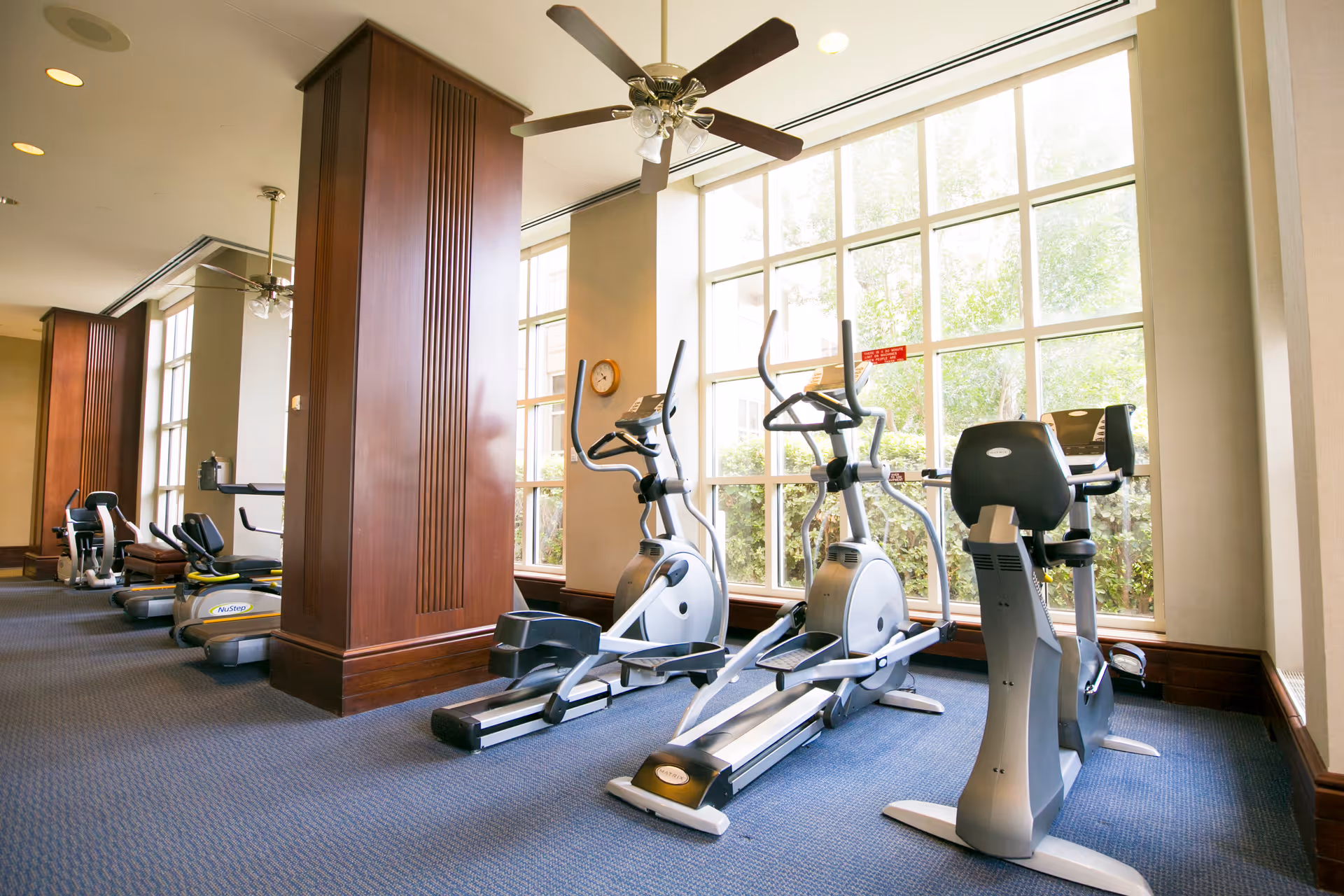 A well-lit fitness room with large windows allowing natural light to fill the space. The room contains several exercise machines including elliptical trainers, stationary bikes, and a NuStep recumbent cross trainer. The walls have wooden paneling and there is a ceiling fan overhead.