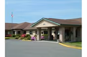 Exterior view of a single-story senior living facility building with a covered entrance, stone pillars, and a flagpole with an American flag. There are landscaped bushes and flowers near the entrance.