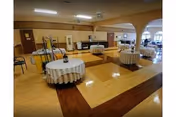 Spacious dining/activity room with round tables draped in white tablecloths, hardwood floor accents, and arched openings to adjacent seating areas.