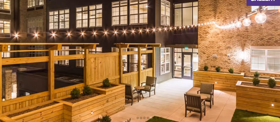 Outdoor patio area at night with wooden planters, small shrubs, string lights hanging overhead, and several chairs and small tables arranged for seating. The surrounding building walls have large windows and brickwork, with a door leading inside.