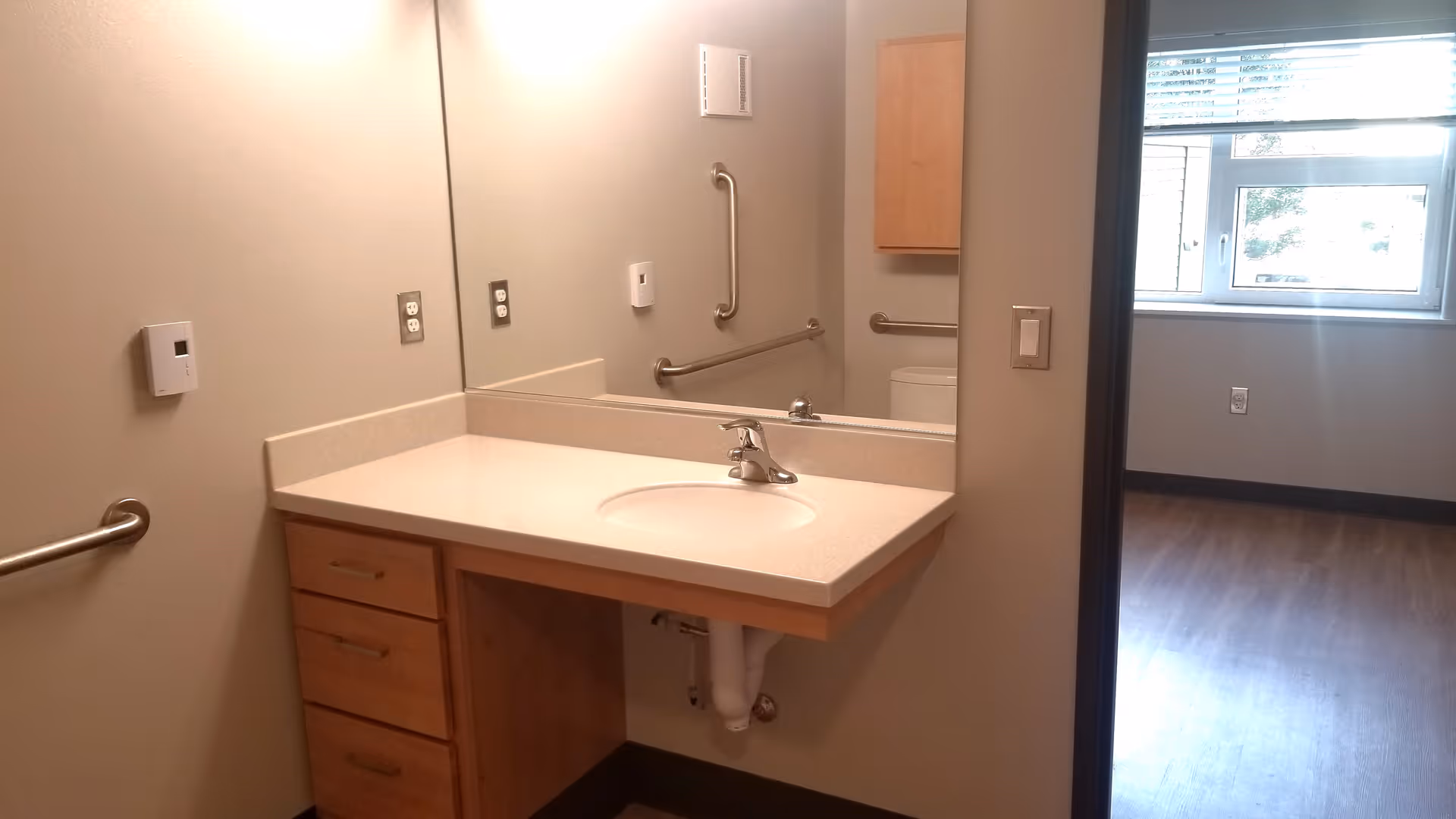 A clean bathroom sink area with a large mirror, beige countertop, wooden drawers, and grab bars on the walls. The image also shows a glimpse of an adjacent room with a window and wooden flooring.