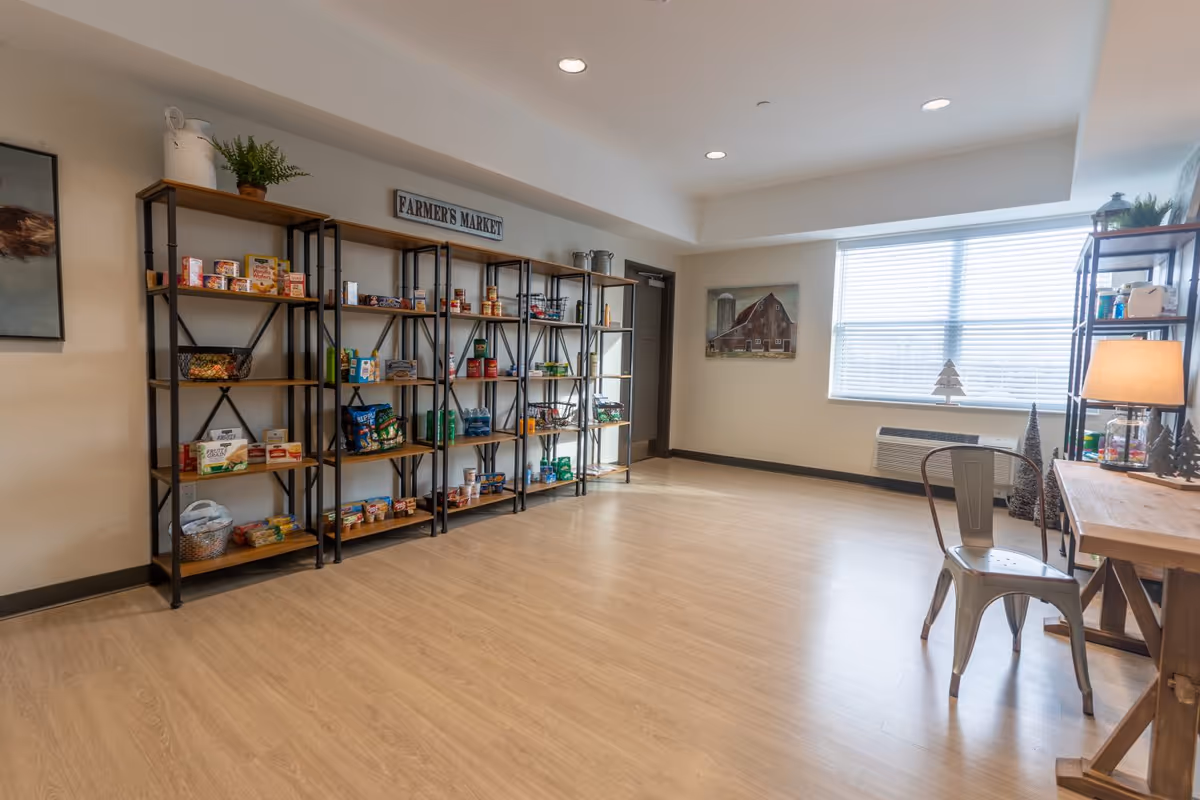 A bright room with wooden flooring featuring metal and wood shelves stocked with various canned and packaged food items under a sign that reads 'FARMER'S MARKET'. There is a metal chair and a wooden table with a lamp on the right side near a large window with blinds. The walls are light-colored with a painting of a barn and some decorative plants.