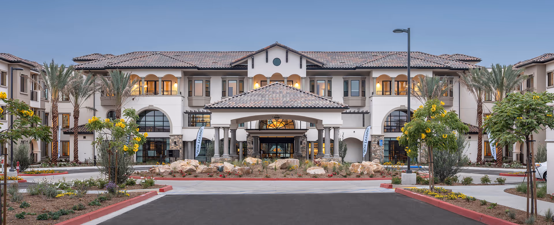 Front exterior of a Mediterranean-style senior living building with a covered porte-cochere, palm trees, and a landscaped driveway.