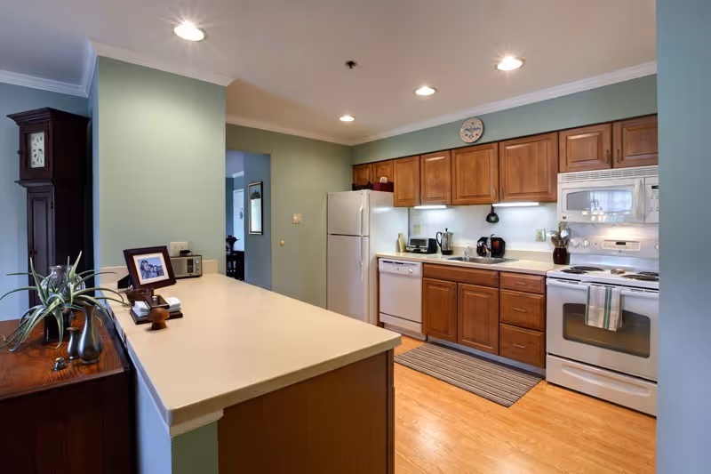 A well-lit kitchen with wooden cabinets, white appliances including a refrigerator, dishwasher, stove, and microwave. The kitchen has a light-colored countertop with a small plant and framed photo on a wooden side table nearby. The floor is wooden, and there is a striped rug in front of the sink area. Recessed ceiling lights illuminate the space.