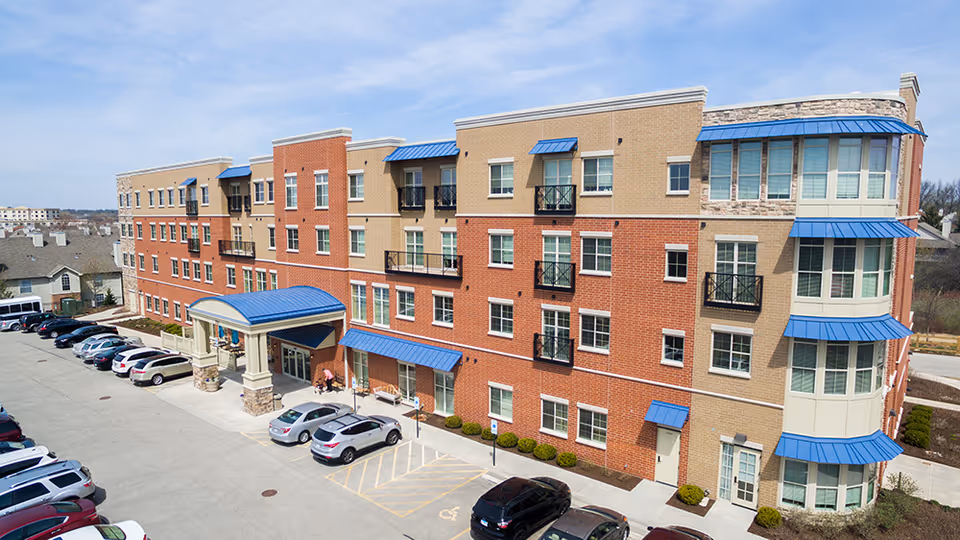 Exterior view of a multi-story assisted living facility building with a brick and beige facade, blue awnings over windows and entrances, a covered main entrance, and a parking lot with several cars parked.