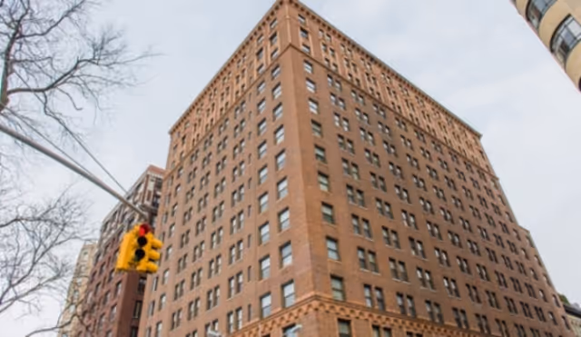 A tall, multi-story brick building with numerous windows, viewed from a low angle against a cloudy sky. A traffic light and some bare tree branches are visible in the foreground.