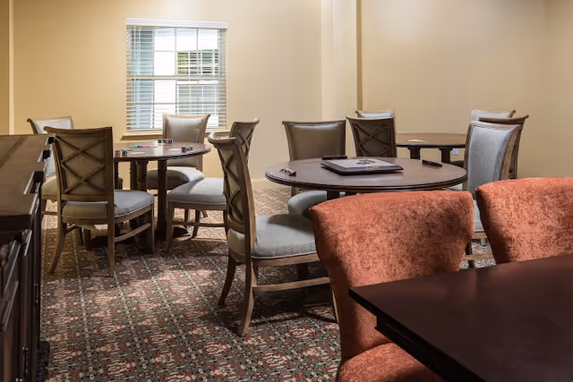 Empty communal dining/activity room with several round wooden tables and upholstered chairs.