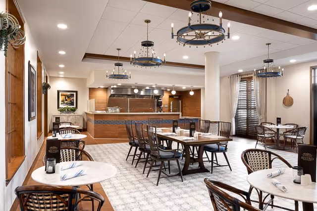 A bright and spacious dining area with multiple round and rectangular tables, each set with napkins and menus. The room features wooden chairs with woven seats, patterned tile flooring, and large windows with curtains allowing natural light. The back of the room has an open kitchen area with stainless steel appliances and a serving counter. Decorative chandeliers hang from the ceiling, adding a warm ambiance.