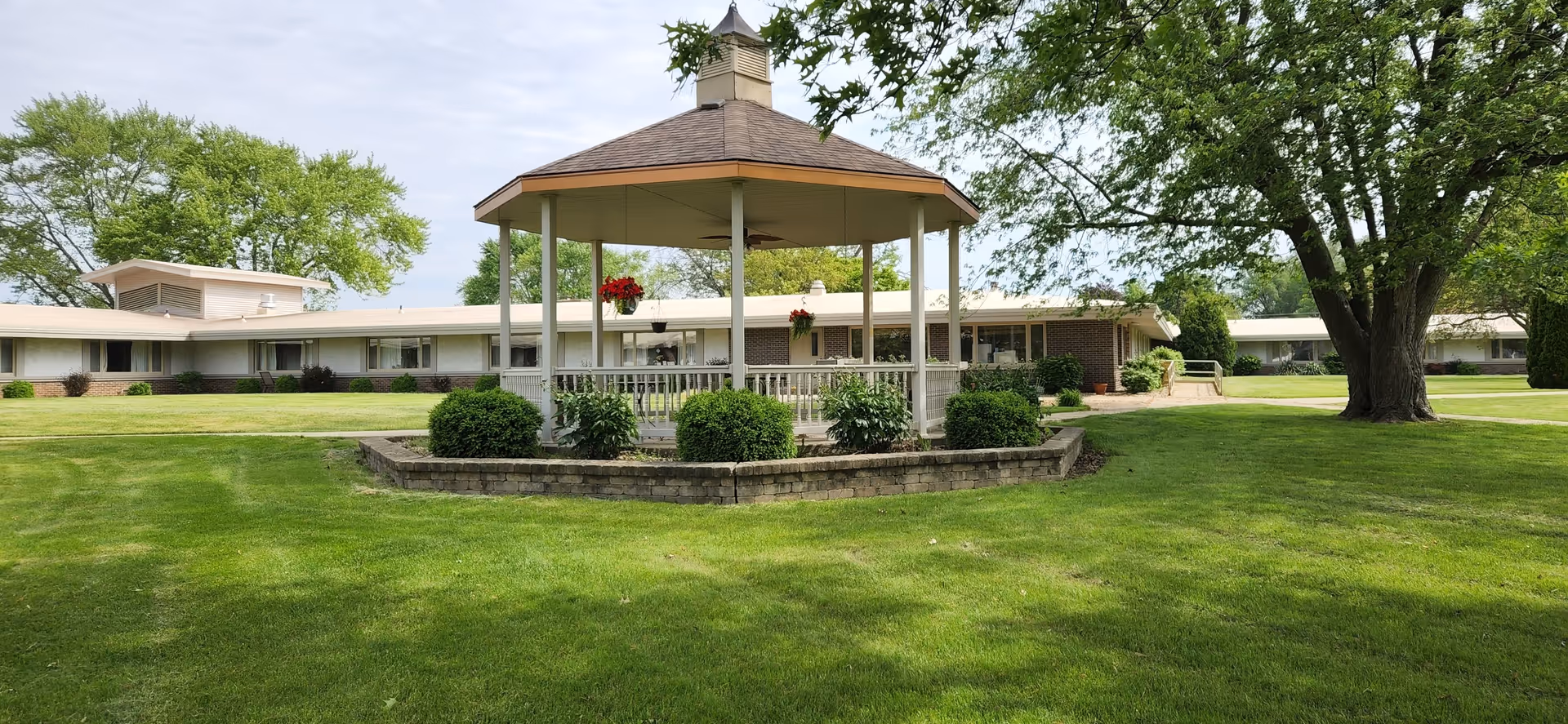 A well-maintained outdoor area of a senior living facility featuring a central gazebo with hanging flower pots, surrounded by green grass, bushes, and large trees. The single-story building with multiple windows is visible in the background under a partly cloudy sky.