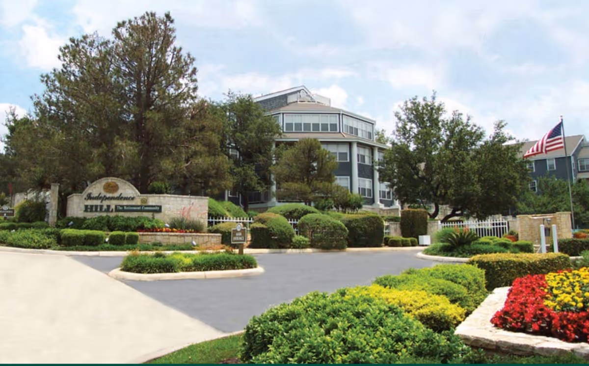 Entrance to Independence Hill Retirement Community featuring a landscaped driveway with trimmed bushes, colorful flower beds, and an American flag. The multi-story building is visible in the background surrounded by trees.