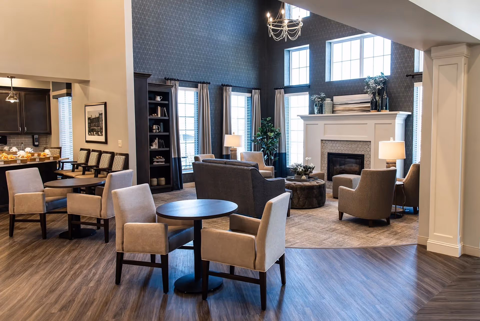 Communal lounge area with round tables, upholstered chairs, a fireplace, and large windows.