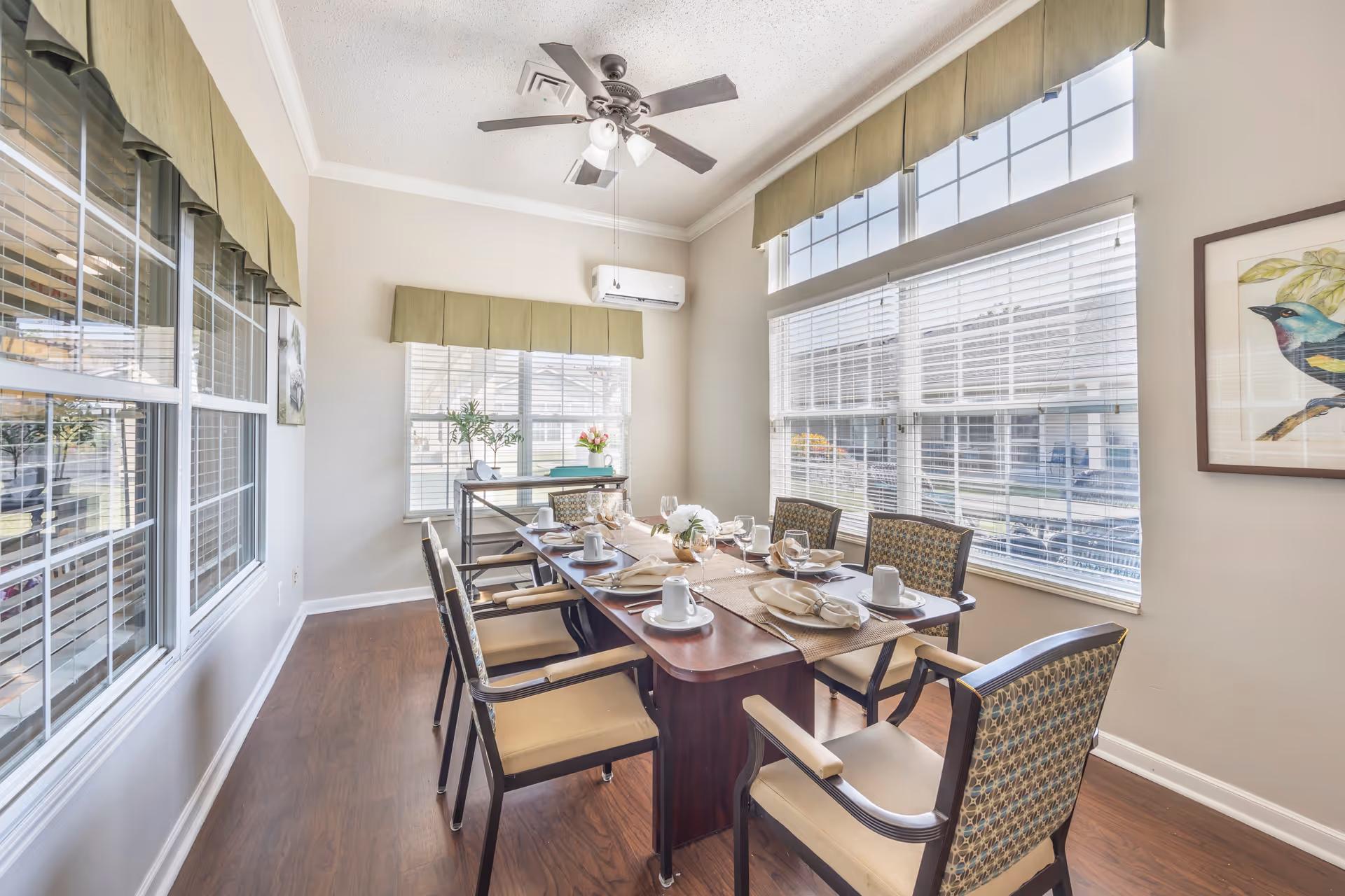 Bright dining room with a long table set for a meal, several chairs, large windows with valances, and a ceiling fan.