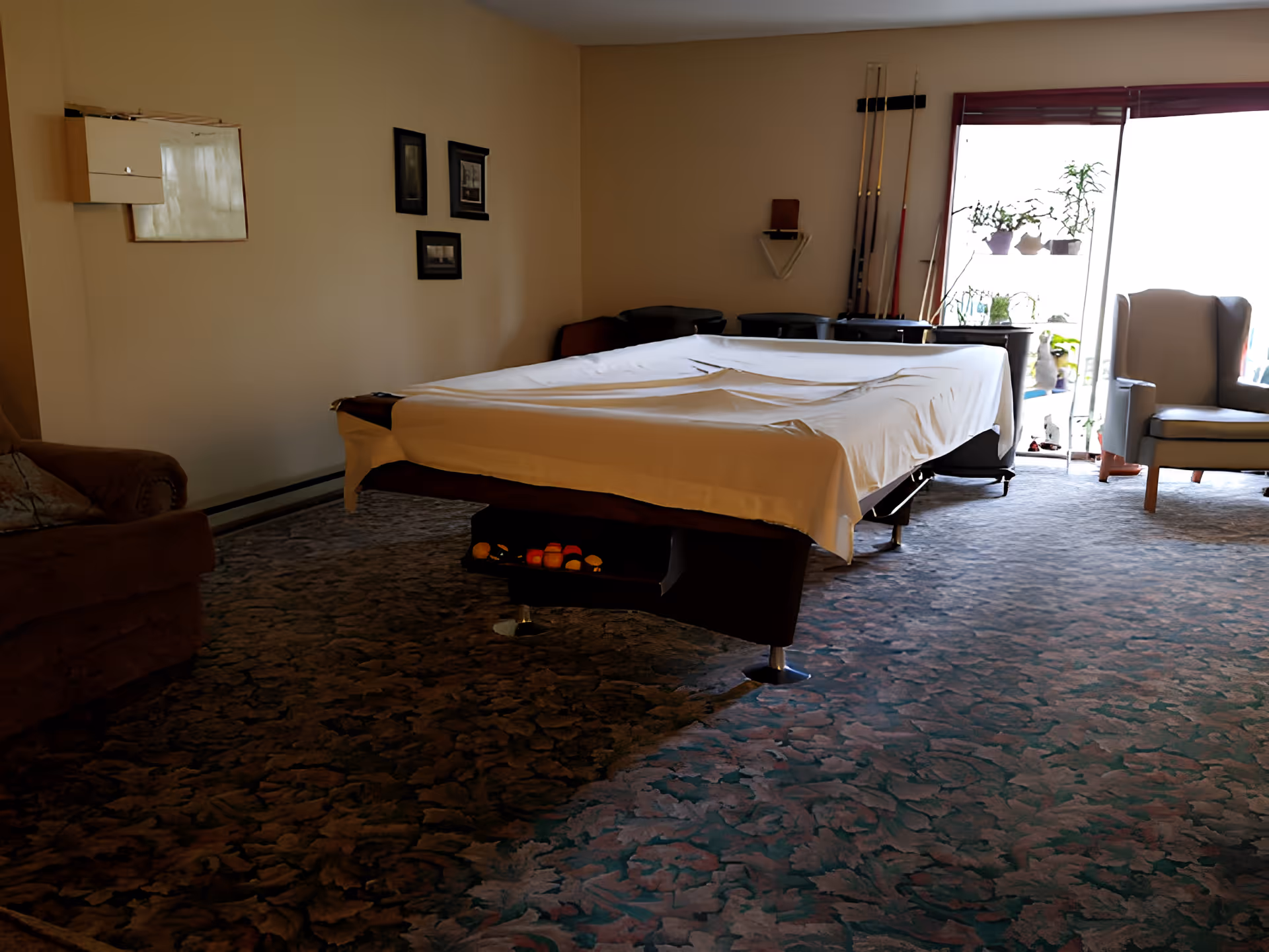 A room with a covered pool table in the center, a brown armchair on the left, a gray wingback chair on the right near a sliding glass door with plants on shelves, and several pool cues mounted on the wall.