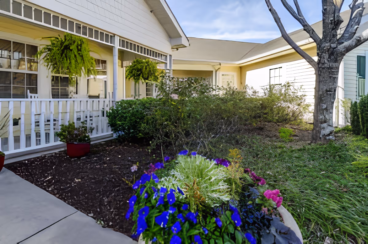 Front porch and garden of a senior living building with hanging ferns, potted flowers, and a walkway.