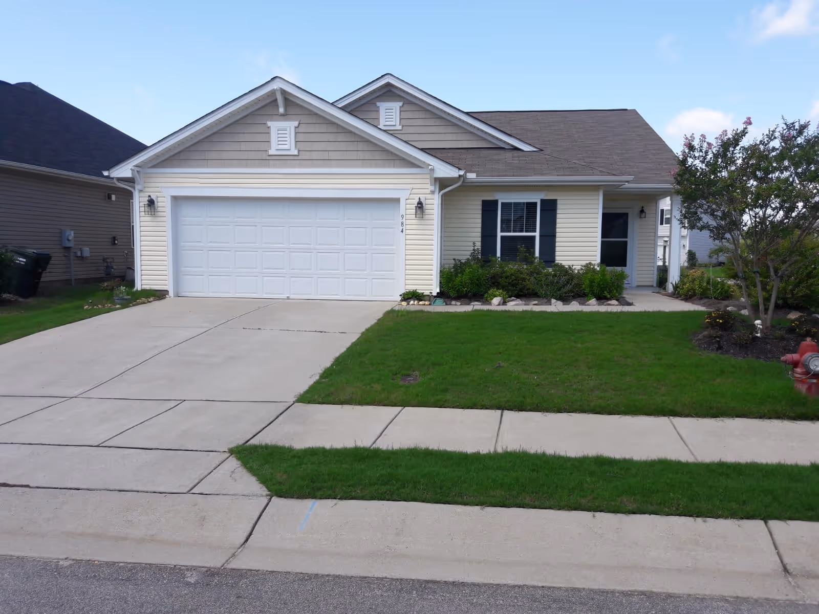 Front exterior view of a single-story house with a two-car garage, beige siding, black shutters, a concrete driveway, green lawn, and a small tree on the right side.