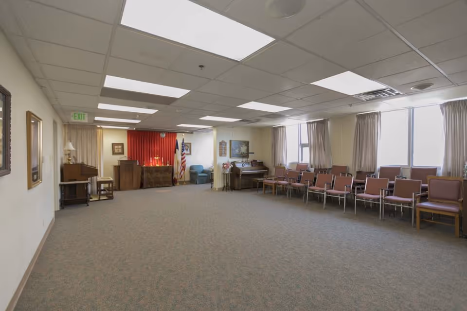 A large room with carpeted floor and fluorescent ceiling lights. Along the right side are multiple rows of chairs with pink cushions, positioned in front of windows with beige curtains. At the far end of the room is a small stage area with a podium, an American flag, and a red curtain backdrop. There is a piano and some framed pictures on the walls, as well as a small table with a lamp on the left side.