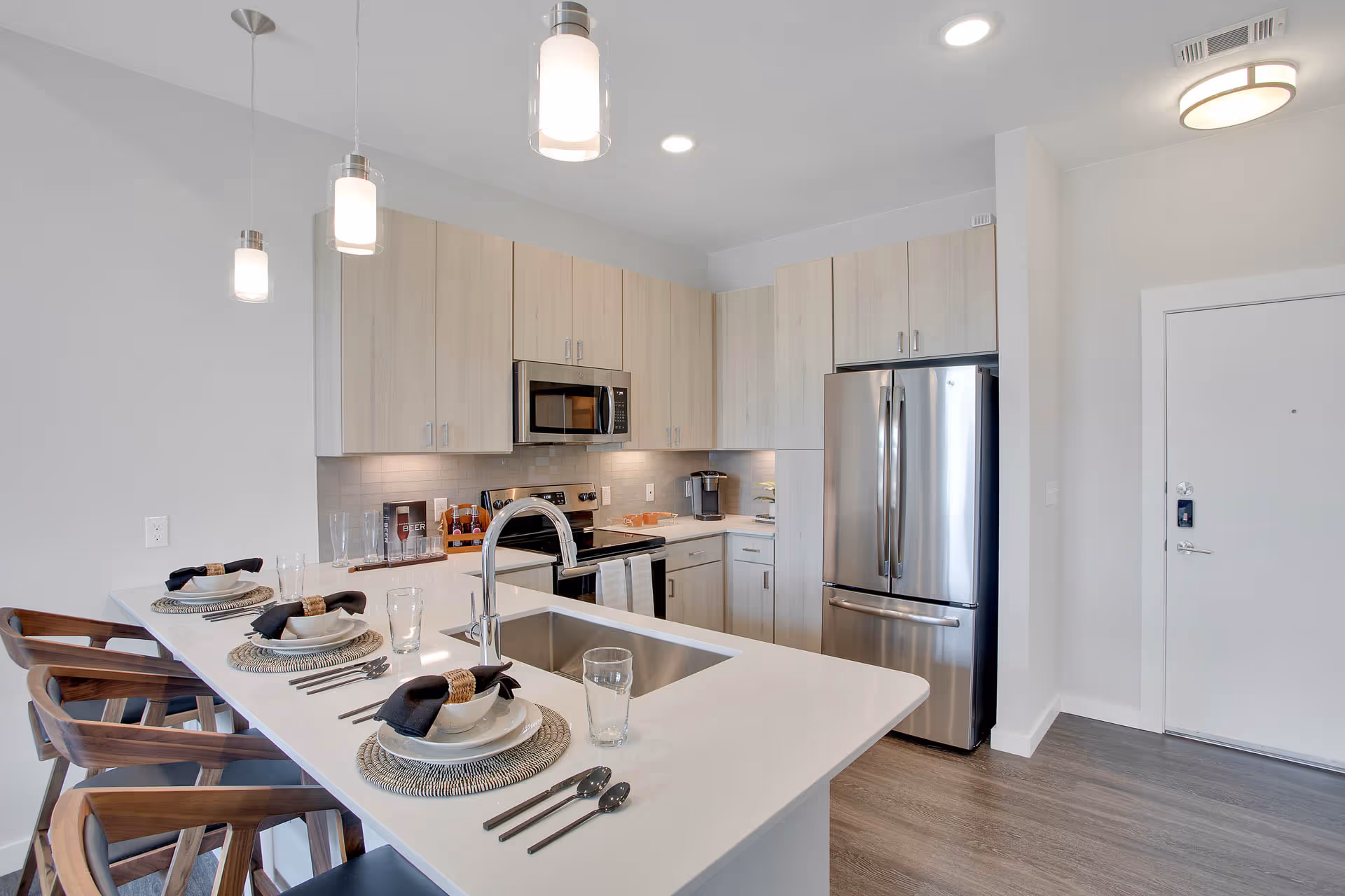 Modern kitchen with light wood cabinets, stainless steel refrigerator, microwave, and stove. A white countertop island with a sink is set with three place settings including plates, bowls, glasses, and cutlery. Three wooden bar stools are positioned at the island. The kitchen has pendant lights and recessed ceiling lights.