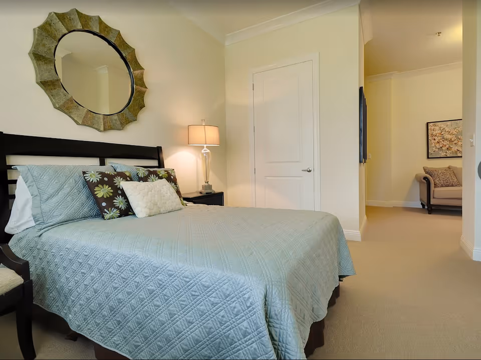 Bedroom with a light-blue quilted bed, decorative pillows, bedside lamp, and round wall mirror above the headboard.