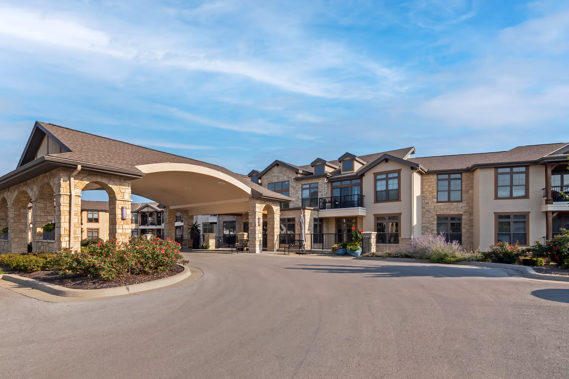 Exterior view of Mission Chateau Senior Living facility showing a large driveway, stone and stucco building with multiple windows, balconies, and a covered entrance with stone pillars. The sky is clear with some clouds.