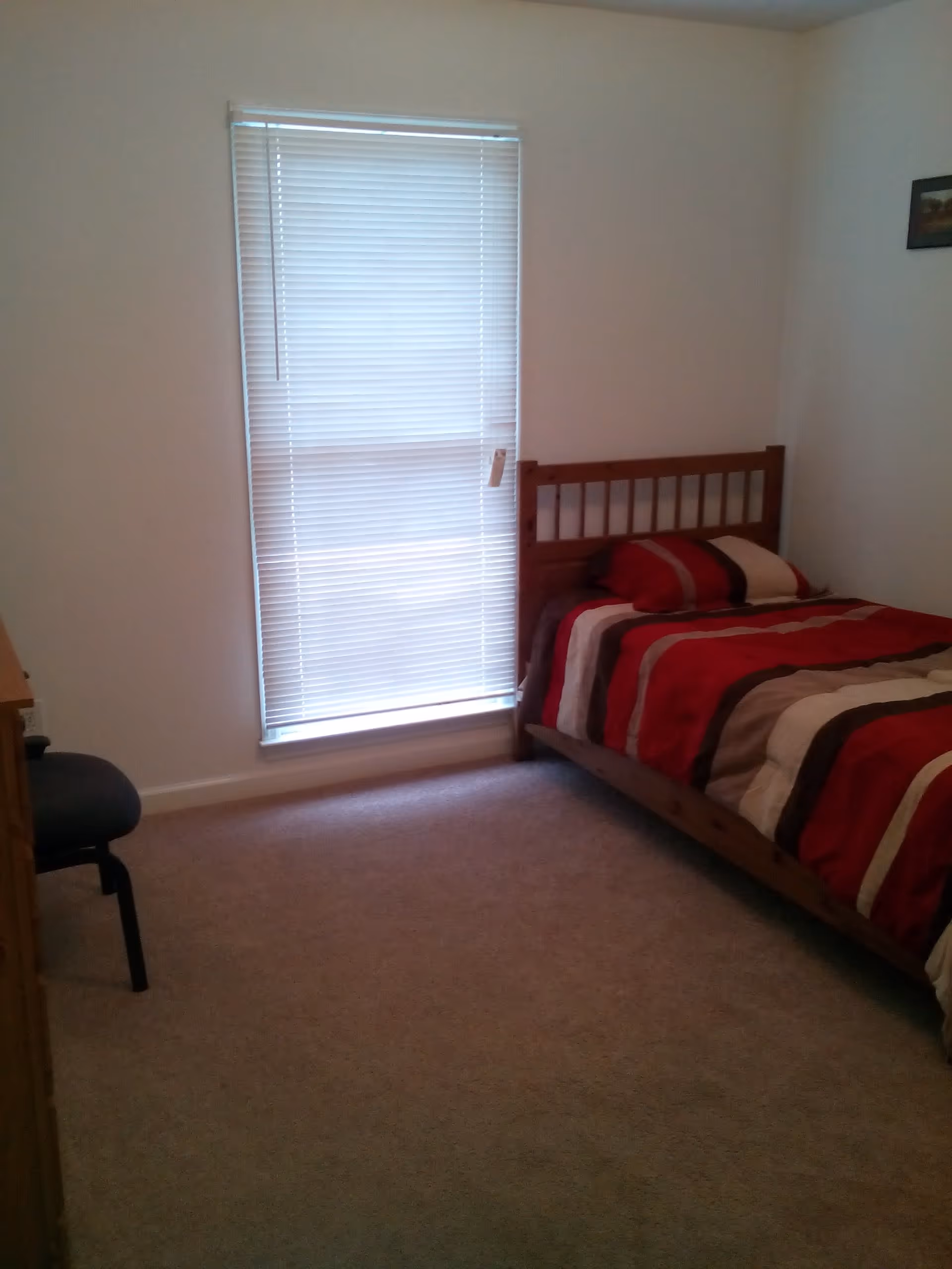 A small bedroom with a single wooden bed against the right wall, covered with a red, brown, and beige striped comforter and matching pillow. There is a window with closed white blinds on the left wall, and a dark chair partially visible in the left foreground. The room has beige carpet and white walls with a small framed picture hanging on the right wall.