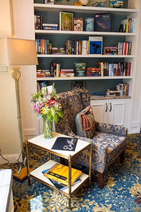 A cozy reading nook featuring a patterned armchair with a decorative pillow, a small side table with a vase of colorful flowers and books, a tall floor lamp, and a built-in bookshelf filled with various books in the background.