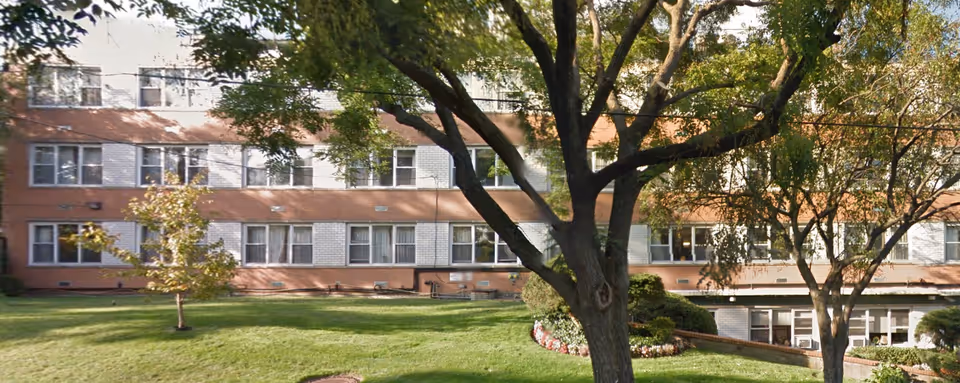 Exterior view of a multi-story brick and white-paneled building with numerous windows, surrounded by green grass, trees, and landscaped bushes.