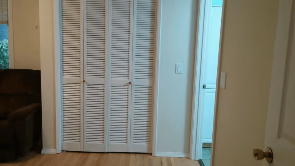 Interior view of a room with light-colored walls and wooden flooring. There is a closed white louvered closet door in the center, a partially visible brown armchair on the left, and an open doorway on the right leading to a room with white cabinetry and tiled floor.