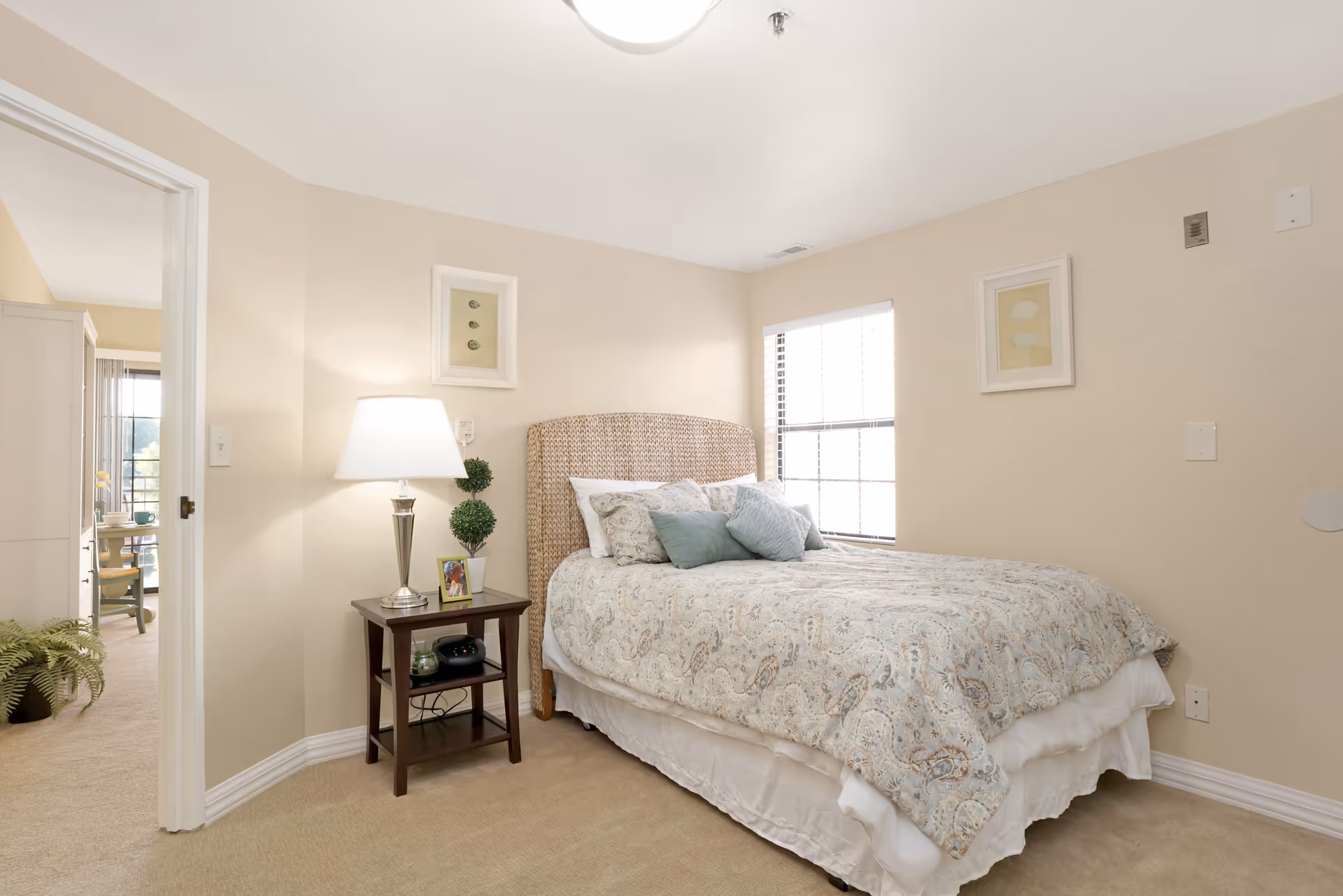 A cozy bedroom with a neatly made bed featuring a patterned comforter and multiple pillows. A wooden nightstand beside the bed holds a lamp, a small plant, and a framed photo. The room has beige walls with two framed artworks and a window letting in natural light. An open door leads to another room with a desk and chair visible.