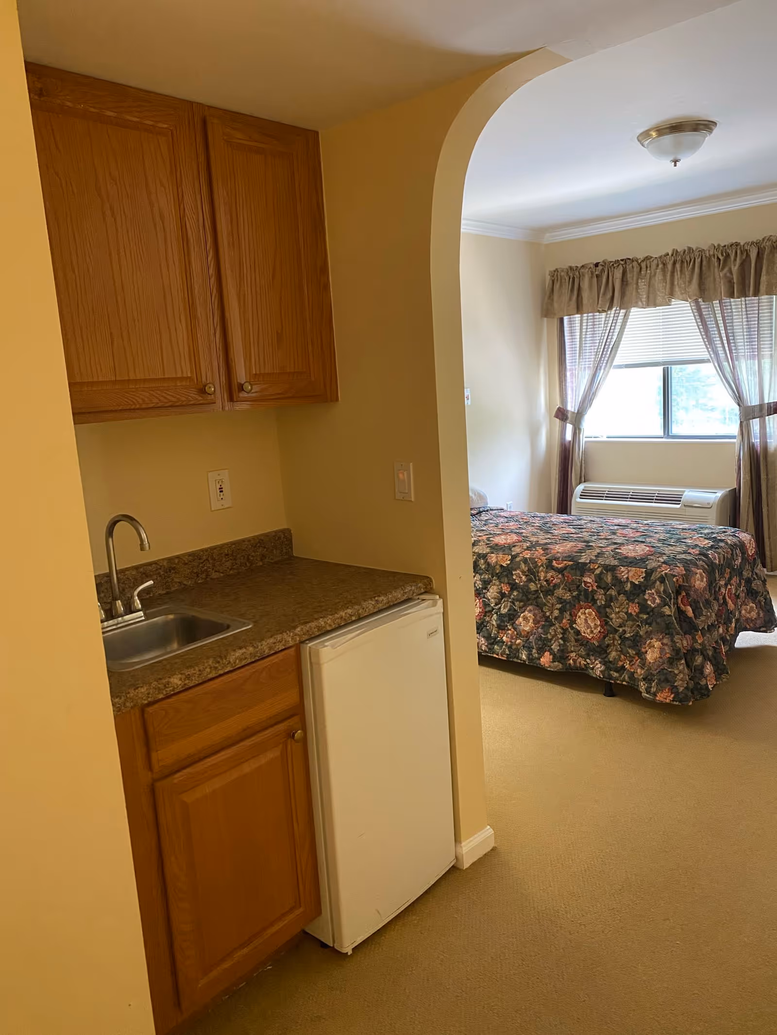 A small kitchenette area with wooden cabinets, a countertop with a sink, and a mini refrigerator. Through an arched doorway, a bedroom is visible with a bed covered in a floral bedspread, a window with curtains, and an air conditioning unit beneath the window.