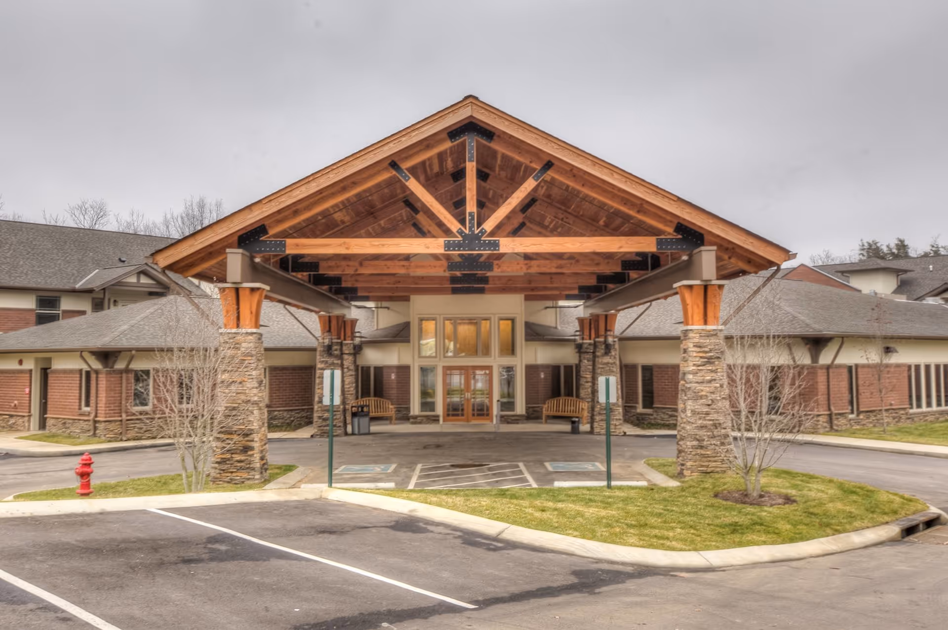 Front exterior view of The Lodge at Natchez Trace featuring a large covered entrance with wooden beams and stone pillars, parking spaces including handicapped spots, and benches near the entrance doors.