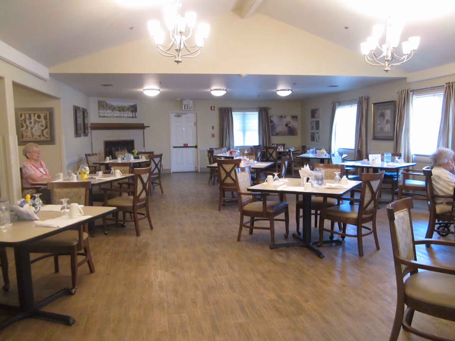 A dining room in an assisted living facility with multiple tables and chairs arranged neatly. The tables are set with cups, glasses, napkins, and pitchers. Two elderly women are seated at separate tables near the walls. The room has wooden flooring, several windows with curtains, framed artwork on the walls, and ceiling lights including chandeliers.