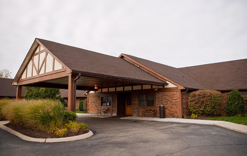 Covered porte-cochere entrance of a single-story brick senior living building with benches and landscaping.