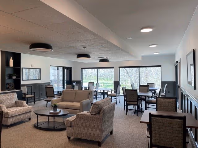 A spacious and well-lit common area with multiple seating arrangements including armchairs, sofas, and tables with chairs. Large windows provide a view of trees and greenery outside. The room has a neutral color palette with beige carpet, gray wainscoting, and white walls. Ceiling lights and modern fixtures illuminate the space.