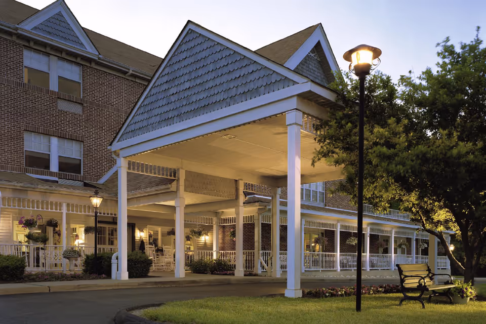 Exterior view of Silver Springs at East Norriton senior living facility during dusk, showing a covered entrance with white pillars, a brick building with multiple windows, a porch with rocking chairs and hanging plants, a lit streetlamp, a bench, and surrounding greenery.