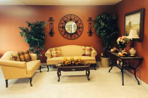 Cozy living room lounge with tan furniture, plants, and decorative wall art inside a facility.