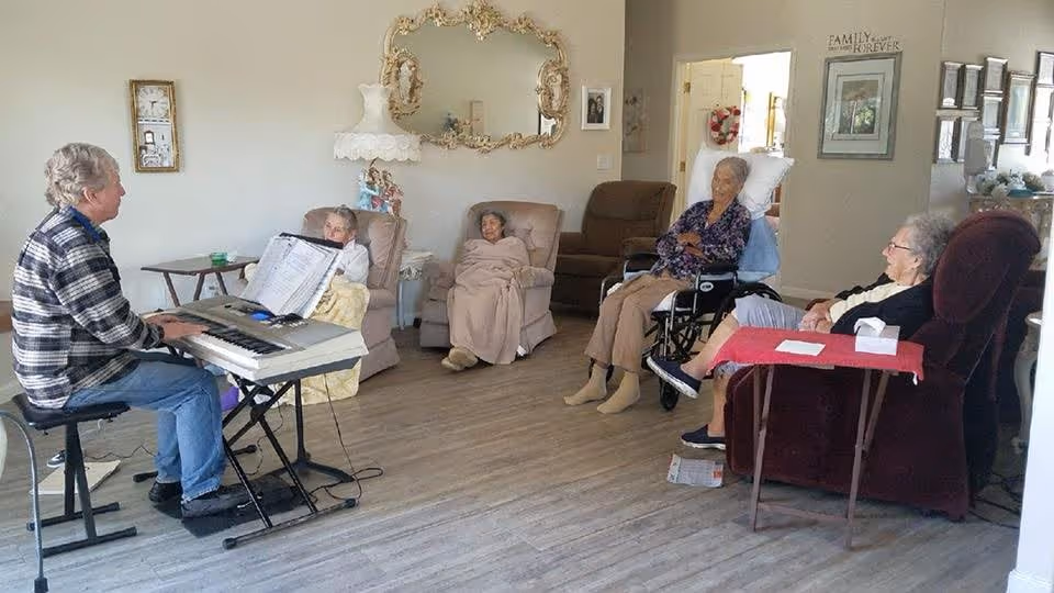 A group of elderly people sitting in a living room area, some in recliners and one in a wheelchair, listening to a man playing an electronic keyboard. The room has light-colored walls, a decorative mirror, framed pictures, and wooden flooring.