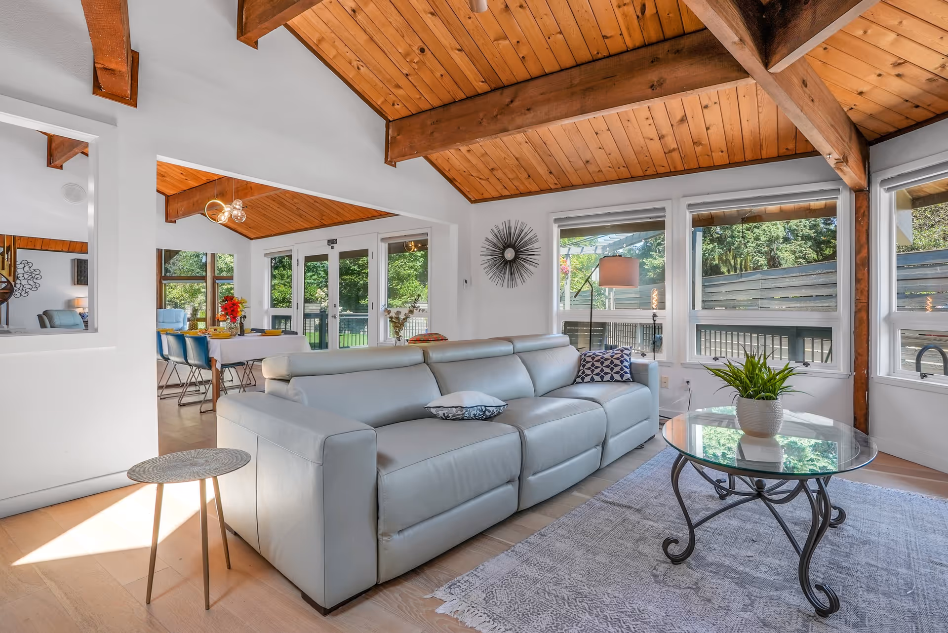A bright living room with a modern light gray leather sofa, a glass coffee table with a plant, and large windows letting in natural light. The ceiling features exposed wooden beams and wood paneling. In the background, a dining area with a table and chairs is visible through an open doorway.