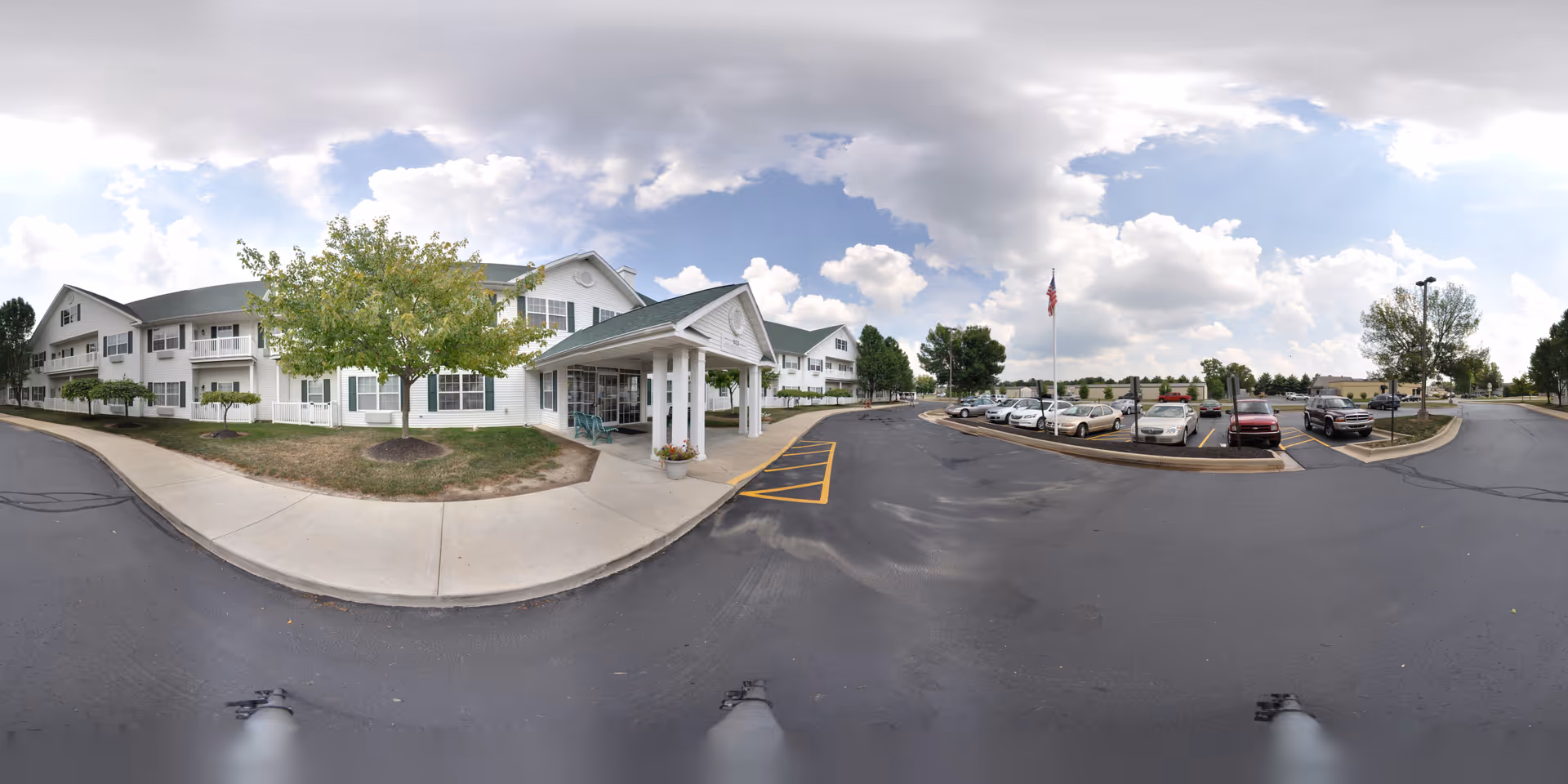 Exterior front of a two-story white senior living facility with a covered entrance, adjacent parking lot, and a flagpole under a cloudy sky.