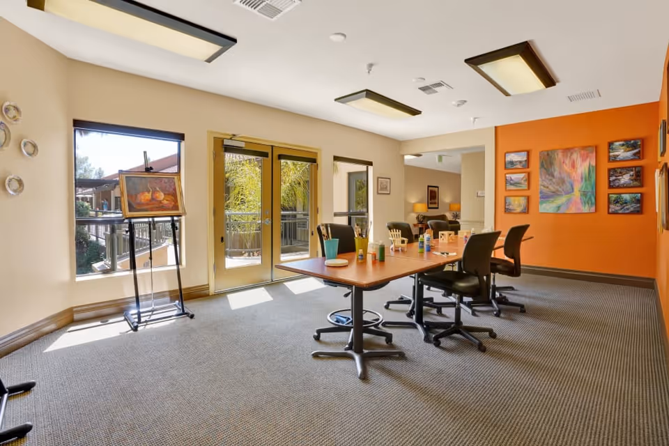 A bright and spacious activity room with a large table surrounded by black office chairs. The table has various art supplies including paint bottles, brushes, and a palette. The room features large windows and glass doors letting in natural light, with an easel holding a painting near the window. The walls are decorated with colorful artwork, including a prominent orange accent wall with multiple framed paintings.