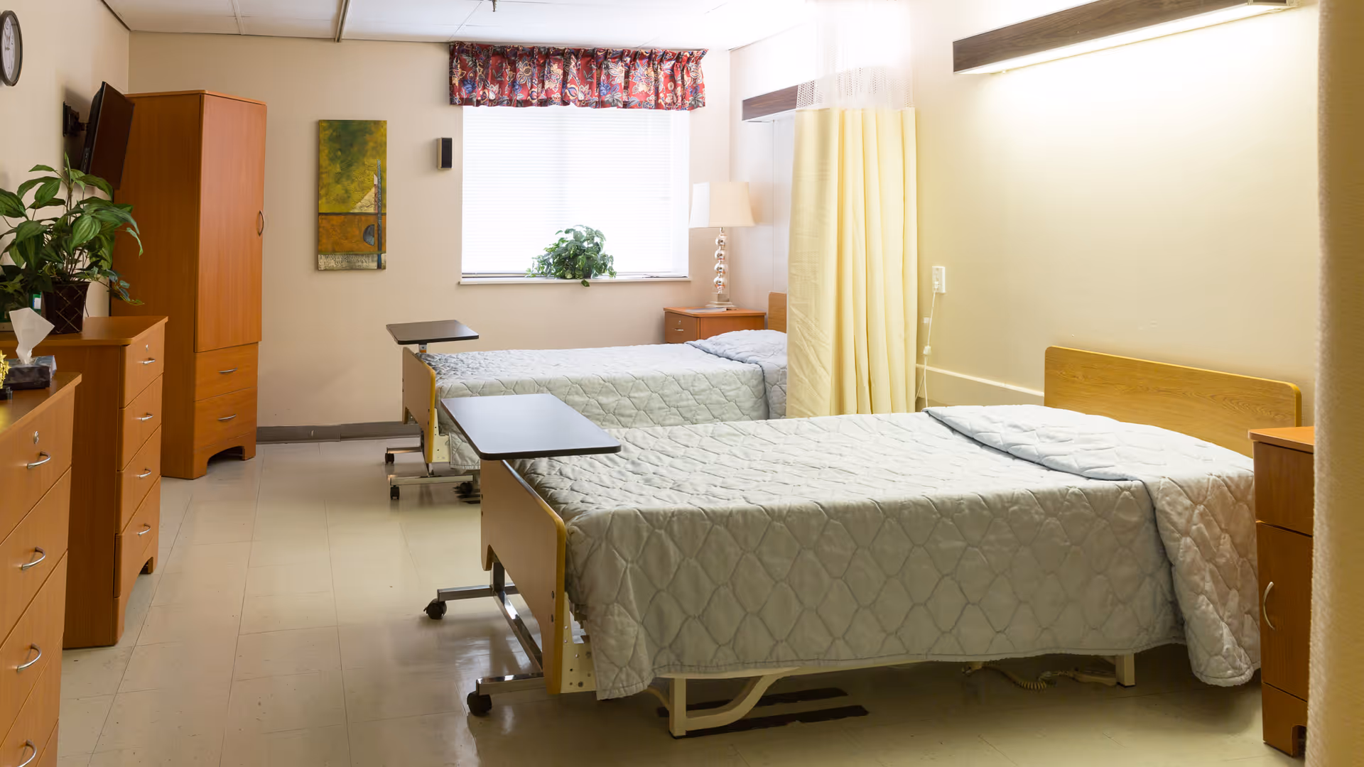 A clean and well-lit room in a rehabilitation and nursing care facility featuring two hospital-style beds with light blue quilted bedspreads. Each bed has an overbed table and wooden headboards. The room includes wooden furniture such as dressers and a wardrobe, a window with a floral valance, a potted plant on the windowsill, a lamp on a nightstand, and a yellow privacy curtain between the beds.
