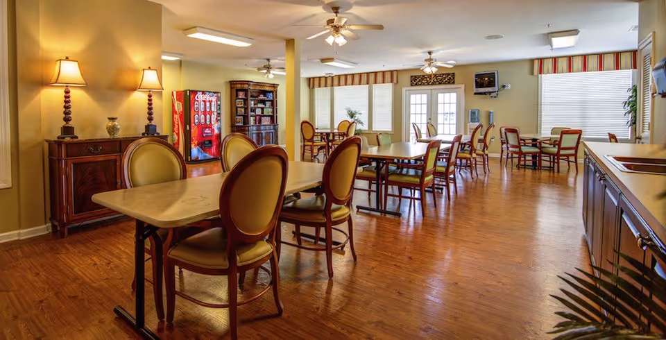 Communal dining room with multiple tables and upholstered chairs, wooden floors, a vending machine and a wall-mounted TV.
